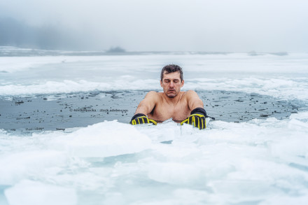 Ein Mann ohne Hemd und mit gelben Handschuhen steht in einem gefrorenen See mit Eisstücken um sich herum. Der Mann hat die Augen geschlossen, und der Hintergrund ist neblig.