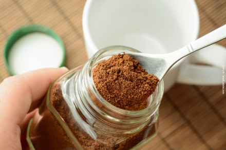 Nahaufnahme eines offenen Glases mit Instantkaffee, wobei ein Löffel mit Kaffeepulver über dem Glas gehalten wird. Im Hintergrund ist eine weiße Tasse und ein grüner Deckel zu sehen.