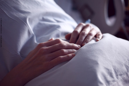 Nahaufnahme von zwei Händen, die auf einer weißen Bettdecke ruhen. Der Hintergrund ist unscharf und deutet auf eine Umgebung im Krankenhaus oder zu Hause hin. Die Hände wirken ruhig und entspannt.