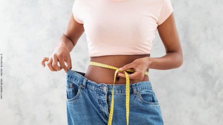 Eine Person trägt eine helle rosa T-shirt und hält ein Maßband um ihre Taille. Die Jeanshose, die sie trägt, ist locker und zeigt, dass sie Gewicht verloren hat.