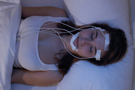 Eine Frau in einem weißen Tanktop liegt in einem Bett und schläft. Ihr Gesicht und Kopf sind mit medizinischen Elektroden und Kabeln verbunden, die mit Klebeband befestigt sind.