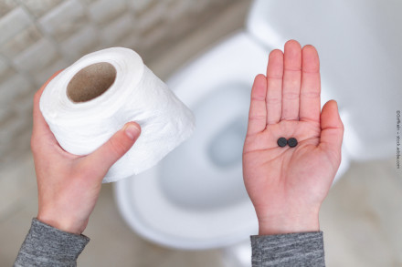 Nahaufnahme von zwei Händen: Die linke Hand hält eine Rolle Toilettenpapier, während die rechte Hand zwei schwarze Tabletten zeigt. Im Hintergrund ist eine Toilettenschüssel zu sehen.