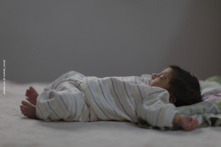Ein Baby in einem gestreiften Schlafanzug liegt auf einem Bett und schläft friedlich. Das Baby hat dunkles Haar und seine Arme und Beine sind ausgestreckt. Der Hintergrund ist verschwommen und zeigt eine ruhige Atmosphäre.