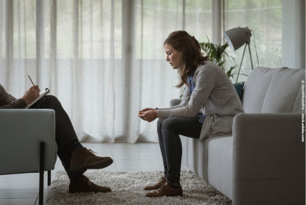 Eine Frau sitzt auf einem Sofa und beugt sich leicht nach vorne. Ihr Gesichtsausdruck wirkt nachdenklich und besorgt. Gegenüber von ihr sitzt eine Person, die ein Notizbuch hält und scheint Notizen zu machen.