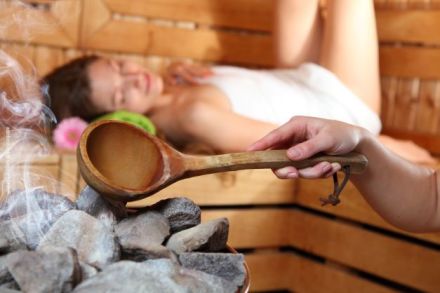Ein entspannter Mensch liegt in einer Sauna. Eine Hand gießt Wasser aus einer Holzschöpfkelle auf heiße Steine, wodurch Dampf entsteht.