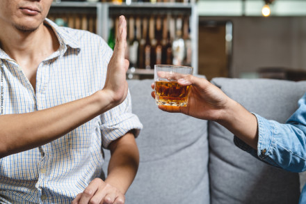 Eine Person in einem karierten Hemd hebt die Hand, um ein alkoholisches Getränk in einem Glas abzulehnen, das von einer anderen Person angeboten wird. Im Hintergrund sind eine Regalwand mit Flaschen und eine gemütliche Raumgestaltung zu sehen.