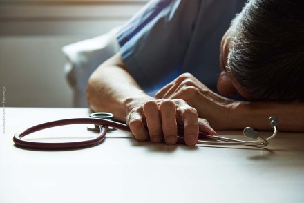 Erschöpfter Arzt in blauer Arbeitskleidung liegt auf einem Tisch und hält in der Hand ein Stethoskop. Sein Kopf ruht auf seinem angewinkelten Arm, während er am Tisch sitzt. Der Raum ist von natürlichem Licht durchflutet.