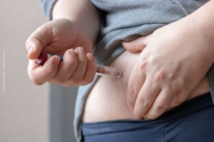 Eine Hand hält eine Spritze und injiziert eine Flüssigkeit in den Bauchbereich einer Person. Die andere Hand zieht die Haut am Bauch leicht zusammen. Die Person trägt ein graues Oberteil und eine dunkelblaue Hose.