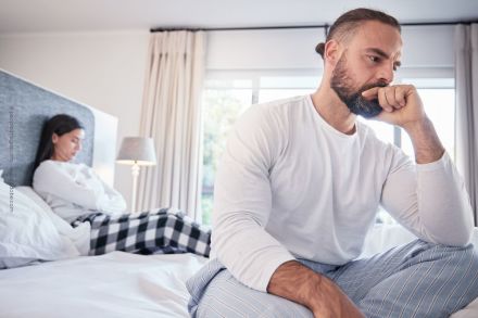 Ein Mann und eine Frau sitzen auf dem Bett und blicken in entgegengesetzte Richtungen. Der Mann im Vordergrund stützt nachdenklich seinen Kopf auf seine Hand, während die Frau im Hintergrund mit verschränkten Armen und gesenktem Kopf sitzt.