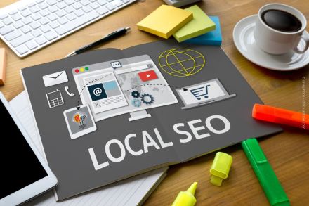 Schreibtisch mit einer Tastatur, einer Tasse Kaffee & Bürobedarf. Im Mittelpunkt steht ein aufgeschlagenes Magazin mit der Aufschrift "LOCAL SEO" und Grafiken zu E-Mail, Onlineshopping, Suchmaschinenoptimierung und Werbung. Notizblöcke und Marker daneben.