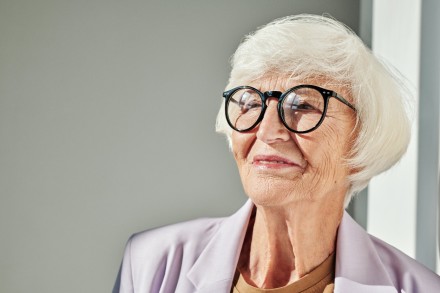 Ältere Frau mit weißem Haar und Brille, schaut nach rechts und lächelt leicht. Sie trägt einen hellen Blazer und steht vor einem neutralen Hintergrund.