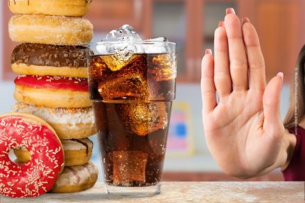 Ein Glas Cola mit Eiswürfeln steht neben einem Stapel bunter Donuts. Eine Hand mit ausgestreckter Handfläche signalisiert "Stopp" oder Ablehnung vor den Donuts und dem Getränk.