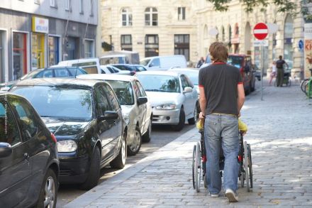 Ein Mann schiebt einen Rollstuhl über einen Bürgersteig auf einer belebten Straße, die von geparkten Autos gesäumt ist. Im Hintergrund sind Geschäfte und mehrstöckige Gebäude zu sehen.