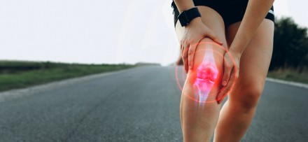 Eine Person hält sich das schmerzende rechte Knie, das von einer digitalen roten Grafik hervorgehoben wird, was auf Gelenkschmerzen oder Arthrose hinweist. Die Person befindet sich auf einer leeren Straße in einer ländlichen Umgebung.