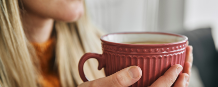 Frau hält eine große rote Tasse in beiden Händen, nah am Gesicht, während sie warme Flüssigkeit genießt. Der Hintergrund ist unscharf, der Fokus liegt auf der Tasse und den Händen. Szene vermittelt Wärme und Entspannung.