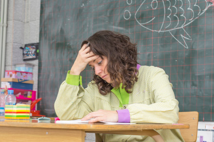 Eine Lehrerin sitzt an ihrem Pult vor der Tafel in einem Klassenzimmer. Sie fasst sich mit der rechten Hand an die Stirn und sieht erschöpft aus.