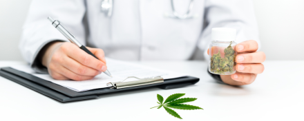 Arzt mit Stethoskop schreibt Rezept und hält Dose mit medizinischem Cannabis. Auf dem Tisch liegt ein Cannabisblatt – Symbolbild für Cannabis auf Rezept.