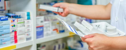 Eine Apothekerin überprüft Medikamente in einem Regal. Sie hält eine Packung in der Hand und prüft die Inhalte, während sie die Medikamente organisiert. Die Regale sind mit verschiedenen Arzneimitteln gut sortiert.