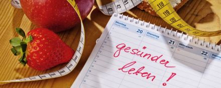 Auf einem Tisch liegen eine Erdbeere, ein Apfel, ein Maßband und ein Kalender mit Aufschrift "gesünder leben".