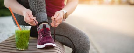 Person in Trainingskleidung bindet sich den Sportschuh. Daneben steht ein grüner Smothie. Symbolbild für einen gesunden Lebenstil