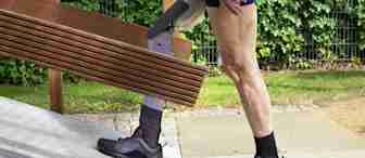A person with a prosthetic leg is carrying wooden planks up a ramp outdoors. They are wearing black socks, black sneakers, and shorts, with a lush green hedge in the background. Their prosthetic limb is visible as they ascend the sloped surface.