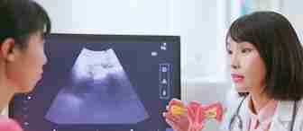 A doctor explains female reproductive health to a patient, holding a model of the uterus and ovaries. An ultrasound image is displayed on the computer screen beside them.
