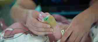 A close-up of an adult hand gently holding the foot of a newborn baby in a hospital. The baby's foot is wrapped in gauze and has medical wires attached, indicating it is receiving medical care. The adult's hand is providing a comforting touch.