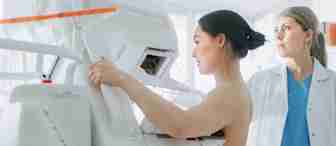 A woman, partially unclothed, undergoes a mammogram in a medical facility. She is positioned beside a mammography machine, with a healthcare professional in a white coat and blue scrubs standing nearby, observing the procedure.