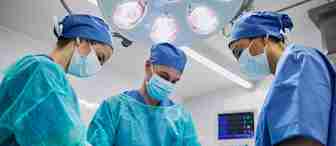 Three surgeons in blue scrubs, masks, and caps perform surgery under bright operating room lights. Their focused attention is on the patient out of view, while medical equipment monitors vital signs in the background.