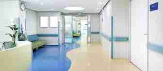 A modern, brightly lit hospital hallway with blue and beige flooring, light green walls, and white doors. There are green cushioned chairs and plants along the left wall. The corridor leads to another section of the hospital through glass doors.
