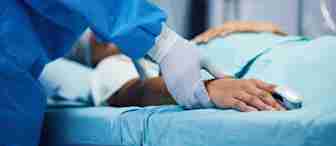 A medical professional wearing blue scrubs and white gloves is holding the hand of a patient lying on a hospital bed. A medical monitor is in the background, displaying vital signs.