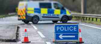 A police van is blocking a road, with bright blue and yellow checkered markings on its side. In front of the van, there are orange traffic cones and a blue "POLICE" sign with a white arrow pointing left, indicating a detour. The background is blurred.