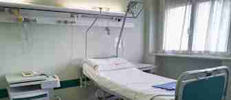 A vacant hospital bed is positioned in a sterile, tidy room. The bed features white linens with a slight pink border, and the room is equipped with medical apparatus. A hospital cart with a phone is next to the bed, and sunlight filters through a window with sheer curtains.