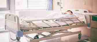 A person lies asleep under a white blanket on a hospital bed in a bright, modern hospital room with wooden walls, medical equipment on the wall, and a large window with blue curtains allowing natural light in.