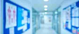 A blurred image of a hospital or clinic hallway with bright overhead lighting. Blue and white informational boards displaying various posters and charts line the walls. The floor and walls are white, contributing to a sterile and clinical environment.