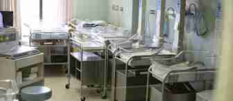 A hospital nursery room with several empty baby bassinets lined up. Each bassinet has a transparent cover and is equipped with basic monitoring devices. The room is softly lit, and curtains cover the windows partially.