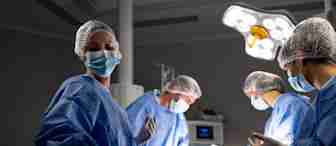 A medical team in surgical uniforms and masks performs a procedure in a brightly lit operating room. One member is adjusting instruments while the others focus on the patient. Medical equipment can be seen in the background.