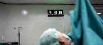 A patient lies on an operating table covered in green surgical drapes, with their face partially visible and a mask over their mouth. In the background, a wall-mounted digital clock displays the time as 8:56:39. Medical equipment and cabinets are also visible.