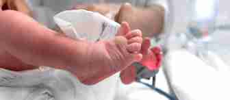 Close-up of a newborn baby's feet in a hospital setting. The baby has atypical cerebral palsy, and a soft light glows on the heel, possibly from a medical device. A healthcare professional's hand is gently holding one of the infant's legs.