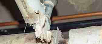 A close-up of a damaged and crumbling asbestos pipe in an industrial setting. The pipe appears to be deteriorating, with exposed sections showing fibrous asbestos material. The surrounding area has other pipes and metallic fixtures visible in the background.