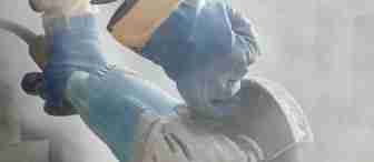 Close-up of a person using an angle grinder to cut a stone. The person is wearing blue protective gloves and there is dust in the air from the cutting process. The angle grinder is blue and appears to be in use.