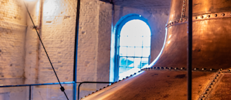 A large copper brewing kettle is seen inside a dimly lit brewery. The room has white brick walls and a large arched window in the background, illuminating the space with natural light. The ceiling is wooden with exposed beams.