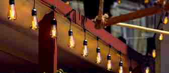 A row of warm, hanging Edison-style light bulbs illuminated under a red wooden pergola. Green plants are in the foreground, and the background contains a blurred structure, all suggesting an outdoor setting, possibly a cozy patio or garden.