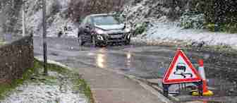 A black car drives cautiously on a wet, winding road with snow-covered sides. Snowflakes fall heavily, and there is a slippery road sign with a traffic cone on the right side, warning of potentially hazardous driving conditions.