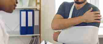 A person with their arm in a sling sits across a desk from a healthcare professional who is writing on a clipboard. Shelves with binders are in the background.
