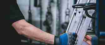A person wearing a black shirt and a blue work glove is holding a metal ladder in a garage or workshop. Out-of-focus tools and equipment are visible in the background, indicating an industrial or mechanical setting.