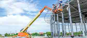Construction workers are standing on an elevated platform attached to a hydraulic lift, working on the exterior frame of a large, partially built steel structure under a clear blue sky. Building materials and land are visible in the foreground.
