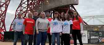 A group of nine people standing in front of a modern, red steel structure. Some wear white shirts with a colorful emblem, while others wear red shirts with "HASAG" printed on them. They are outdoors on a cloudy day, with buildings and signs in the background.