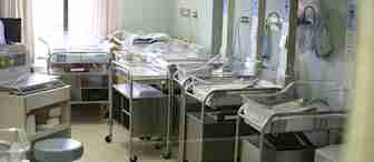A hospital nursery room with multiple empty baby cribs lined up. Each crib is equipped with a monitoring device. The room has pastel green walls, curtains, and various medical equipment. There are no people present.