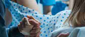A person in a hospital gown with blue polka dots is lying in a hospital bed, gripping another person's hand tightly. Medical professionals wearing blue gloves are visible in the background, suggesting that the person is in labor.